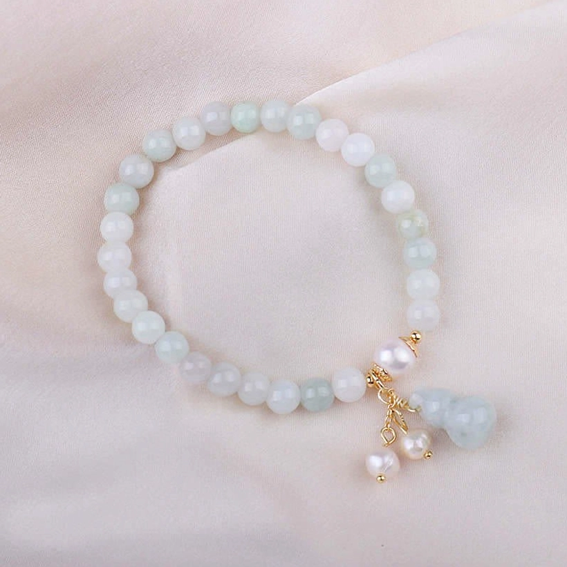 Jade pearl gourd luck bracelet flat lay on soft light fabric surface displaying milky pale green jade beads, gold fittings, and pearl gourd pendant