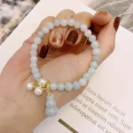 Natural jade pearl gourd prosperity bracelet with pale green jade beads, gold hardware, and dangling pearl gourd charm held in hand on book background