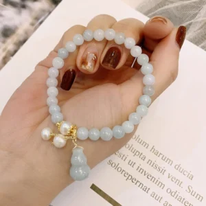 Natural jade pearl gourd prosperity bracelet with pale green jade beads, gold hardware, and dangling pearl gourd charm held in hand on book background