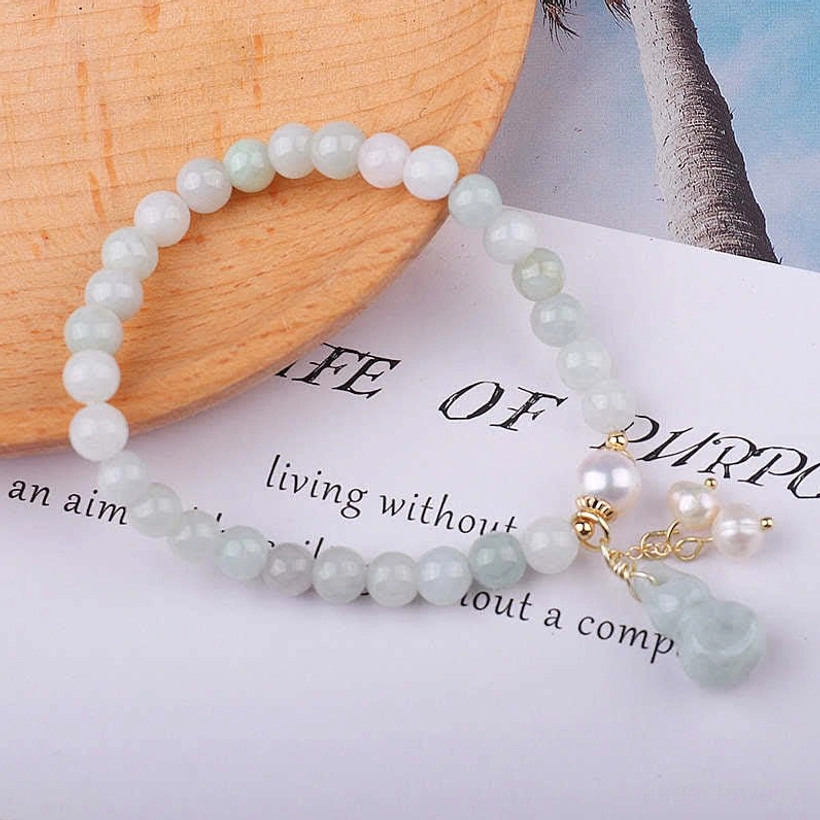 Jade pearl gourd prosperity bracelet flat lay with pale green jade beads, dangling pearl gourd charm, gold accents on book with wooden coaster and palm backdrop