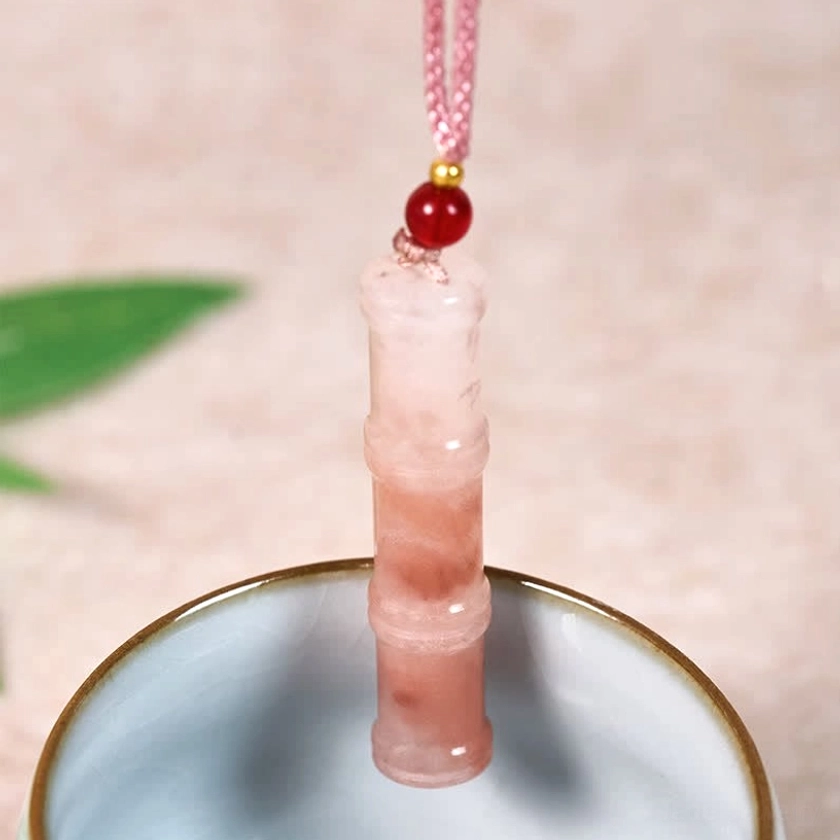 Vertical display of translucent pink quartzite jade bamboo pendant necklace suspended over ceramic bowl with botanical elements, showcasing stone clarity and light transmission