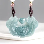 Natural jade peony ruyi lock pendant necklace carved with floral motifs, front view displayed on white ceramic platform, bluish jade prosperity pendant