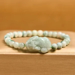 Close-up flat lay view of jade Pixiu beaded bracelet on brown wooden surface featuring larger carved Pixiu focal piece with gradient jade stone tones