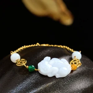 Natural jade Pixiu copper coin luck bracelet with white jade charm, gold chain, and green orange bead accents on dark stone background