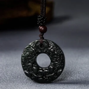 Nanyang black-green jade Pixiu peace buckle pendant with relief carved Pixiu design, hanging against dark gray fabric background with woven rope