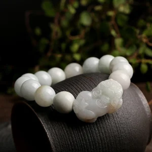 Milky white jade beads with white carved Pixiu ornament on dark cylindrical display stand with green foliage background