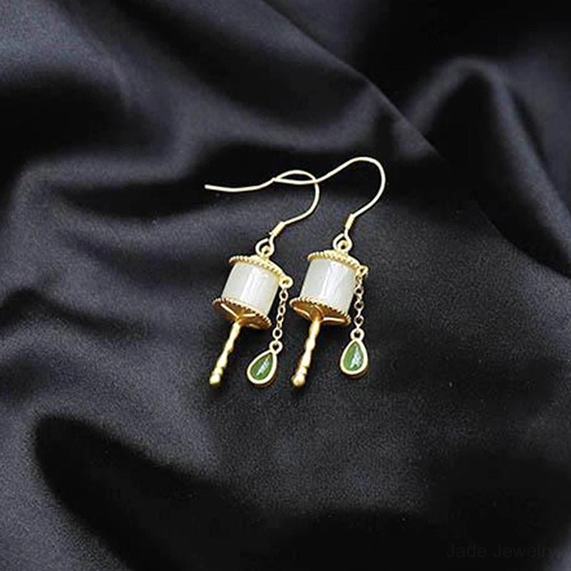 Jade prayer wheel drop earrings displayed on black velvet fabric, showing gold ear hooks, milky white jade tube, and green teardrop pendants with minimalist luxury aesthetic