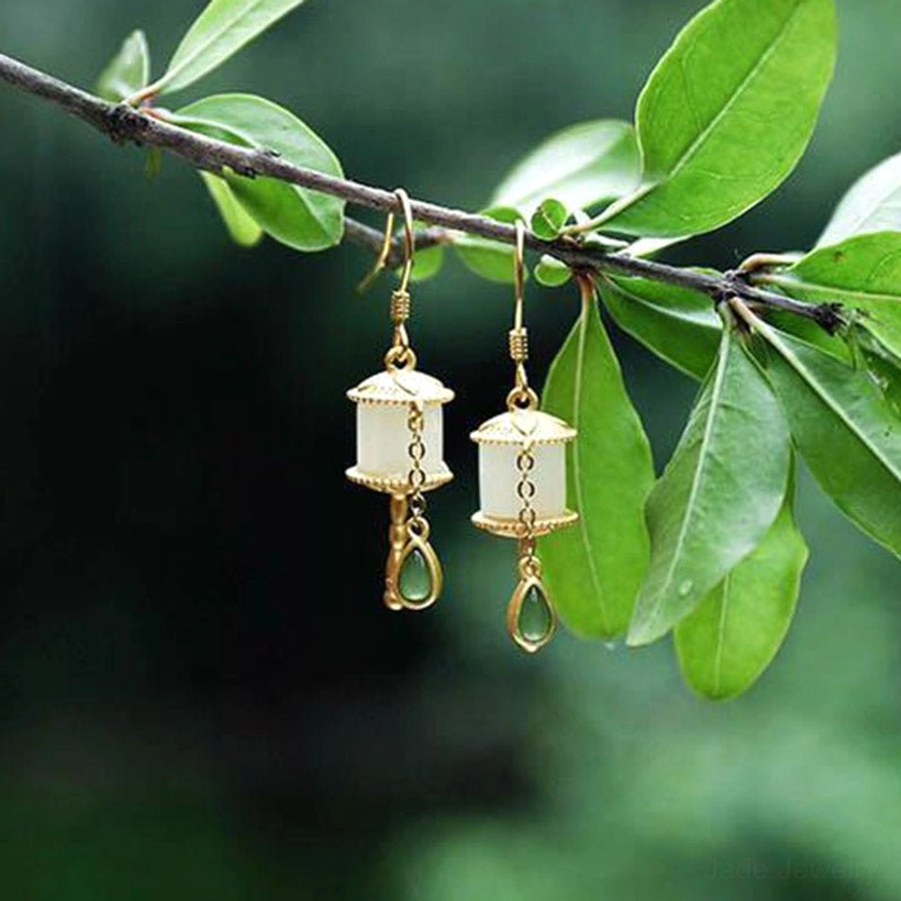 Eye-level view of jade prayer wheel drop earrings hanging from tree branch, showcasing traditional oriental design with gold metal structure and green teardrop pendant
