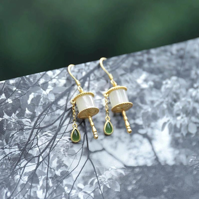 Pair of jade prayer wheel drop earrings on forest pattern background, featuring sterling silver accents, milky white jade tubes, and green teardrop charms