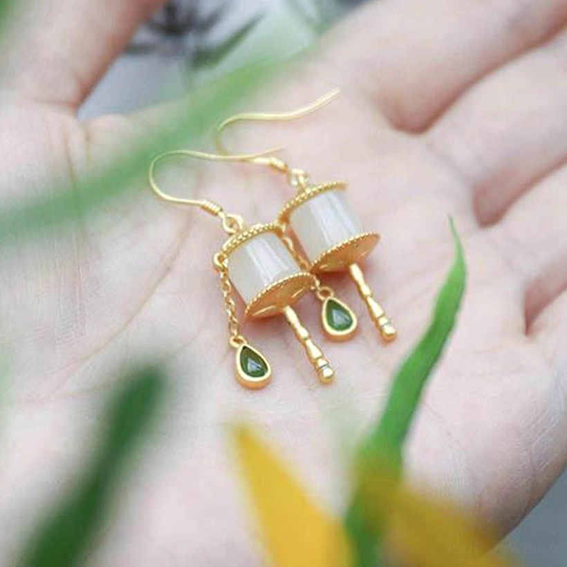 Jade prayer wheel drop earrings held in palm, displaying sterling silver hooks, milky white jade beads, and green teardrop charms against outdoor plant background