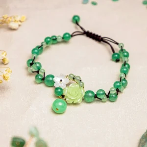 Green jade braided anklet with black cord and floral charms displayed on tabletop with dried florals