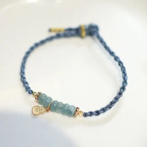 Jade prosperity bracelet with gold metal pendants and dark blue woven cord displayed on white ceramic plate