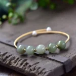 Green jade beaded bracelet with 14K gold frame displayed on vintage wooden surface with green foliage, featuring gradient jade beads and white pearl accents