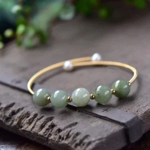 Green jade beaded bracelet with 14K gold frame displayed on vintage wooden surface with green foliage, featuring gradient jade beads and white pearl accents