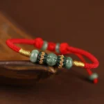 Red string jade prosperity bracelet with gold twisted thread accents, displayed on wooden surface from side angle