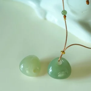 Multiple natural jade prosperity pendant variations in celadon and light green shades showing color range options with gold-tone accent beads on braided cord