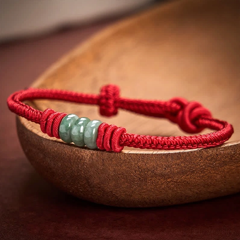 Single red string jade bracelet with light green jade beads and sliding knot resting on wooden bowl