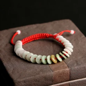 Natural jade round beads with red protective string bracelet displayed on vintage dark box, showing mixed-tone jade discs and braided red cord details