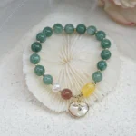 Green jade Ruyi lock bracelet with sea shell charm displayed on white seashell and sand background - top view flat lay