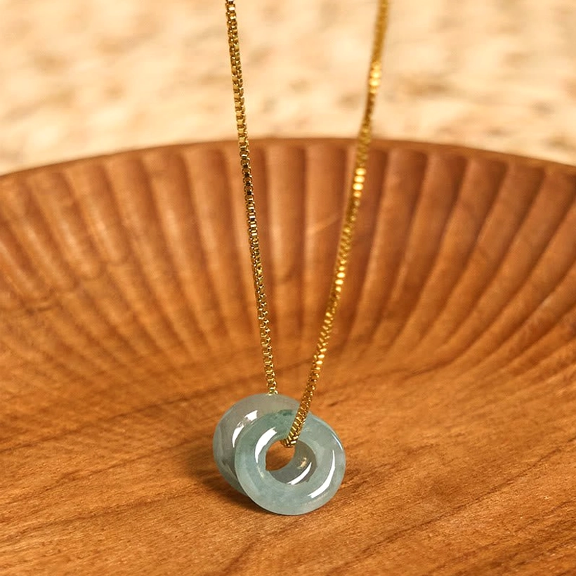 Single natural jade peace buckle pendant necklace with gold chain on wooden tray, front overhead view