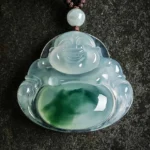 Laughing Buddha Maitreya cyan jade pendant necklace in flat lay composition with braided cotton cord and jade bead accent on dark textured stone surface, showing translucent ice blue jade with green belly patch