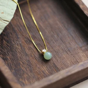Light green jade prosperity pendant necklace with 925 sterling silver gold chain on dark wooden tray, overhead view with natural leaf detail