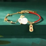 Lunar Rabbit white jade red string bracelet with fortune charm displayed flat lay on dark green background, showing gold-plated hardware and coin-shaped pendant