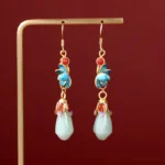 Magnolia jade drop dangle earrings with blue enamel flowers, gold ear hooks, and light green jade buds displayed on metal rack