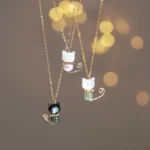 Three handmade Maneki-neko lucky cat necklaces with gold-plated chains and jade stone accents displayed hanging against warm bokeh background