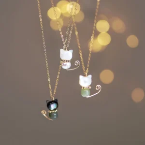Three handmade Maneki-neko lucky cat necklaces with gold-plated chains and jade stone accents displayed hanging against warm bokeh background