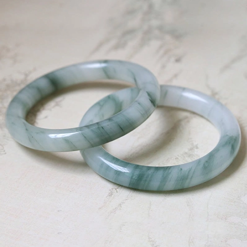 Pair of matching natural jade bangles with dark green flower patterns displayed on light table surface, flat lay top view showing polished finish
