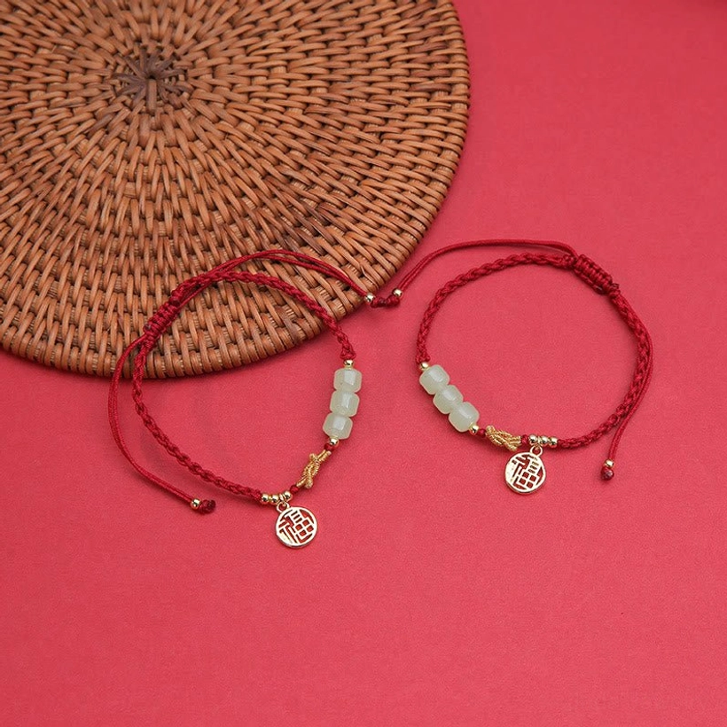 Pair of matching handcrafted jade Fu bracelets with red rope and gold charms displayed together on rattan mat