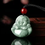 Natural green jade Laughing Buddha Maitreya pendant with hand-carved smiling face, black adjustable braided cord with red glass bead accent, front oblique view on wooden display stand