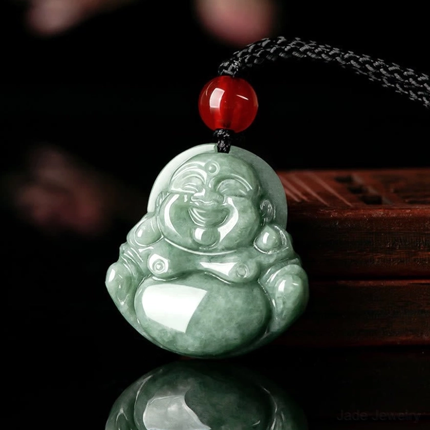 Natural green jade Laughing Buddha Maitreya pendant with hand-carved smiling face, black adjustable braided cord with red glass bead accent, front oblique view on wooden display stand
