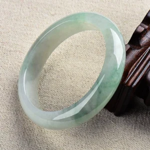 Natural jade bangle bracelet with light green and white tones displayed on wooden stand, showing smooth polished finish and natural vein patterns
