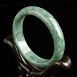 Natural green jade bangle bracelet displayed on wooden stand, showing polished mottled jade texture from left oblique angle