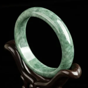 Natural green jade bangle bracelet displayed on wooden stand, showing polished mottled jade texture from left oblique angle