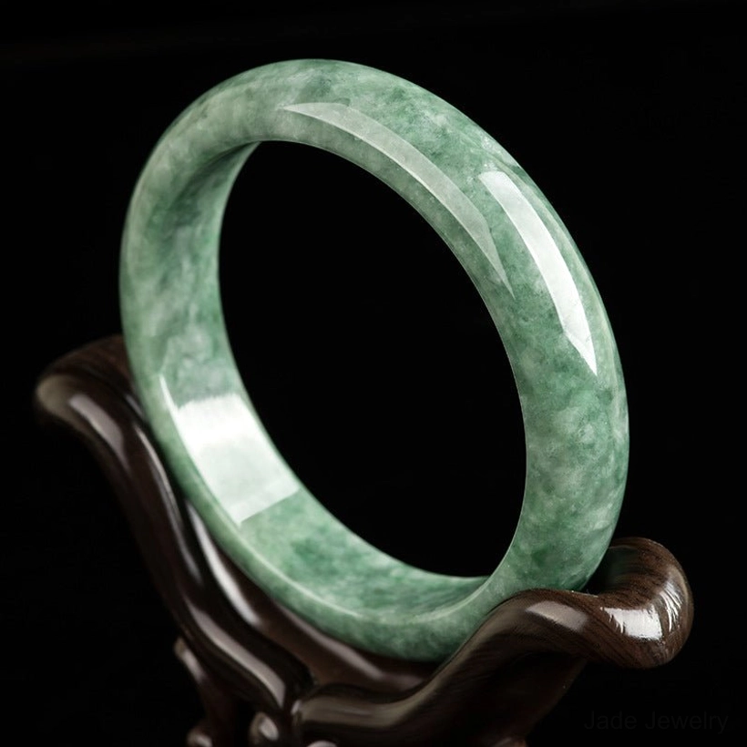 Natural green jade bangle bracelet displayed on wooden stand, showing polished mottled jade texture from left oblique angle