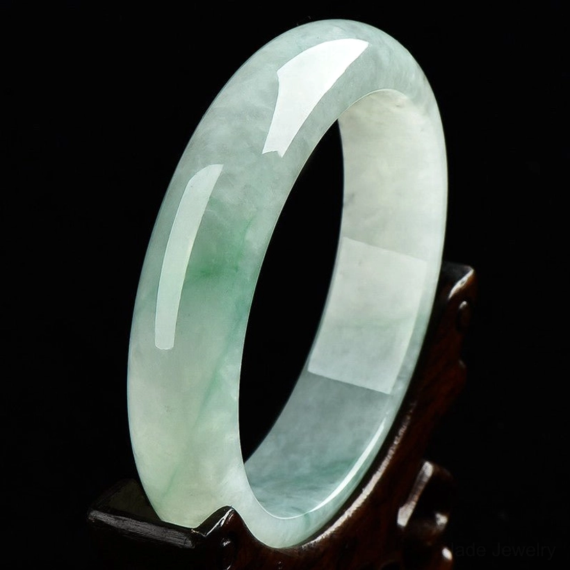 Pale green translucent jade bangle with natural jadeite inclusions, mounted on carved wooden display stand against dark background, highlighting jade's luminous quality
