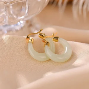 Close-up of light natural jade drop earrings with 925 sterling silver gold-plated lever-back closure on elegant satin fabric with glassware backdrop