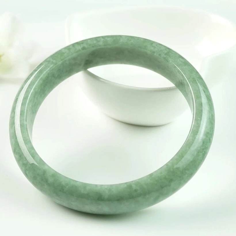 Natural green jade luck bangle on white background with ceramic bowl, left oblique close-up showing authentic jade patterns