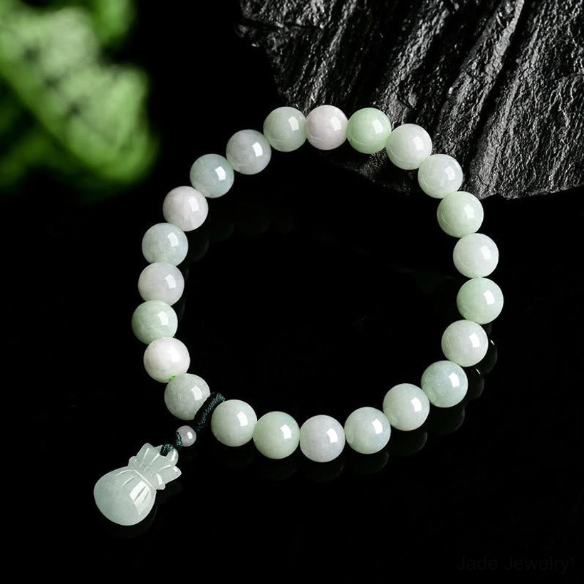 Jade bracelet with carved money bag charm photographed on dark background with decorative stone and foliage, pale green beads detail
