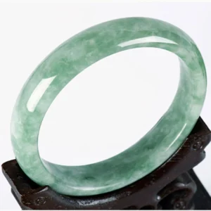 Natural jade prosperity bangle bracelet with light green mottled texture displayed on carved wooden holder, three-quarter top-down view showing authentic jade craftsmanship