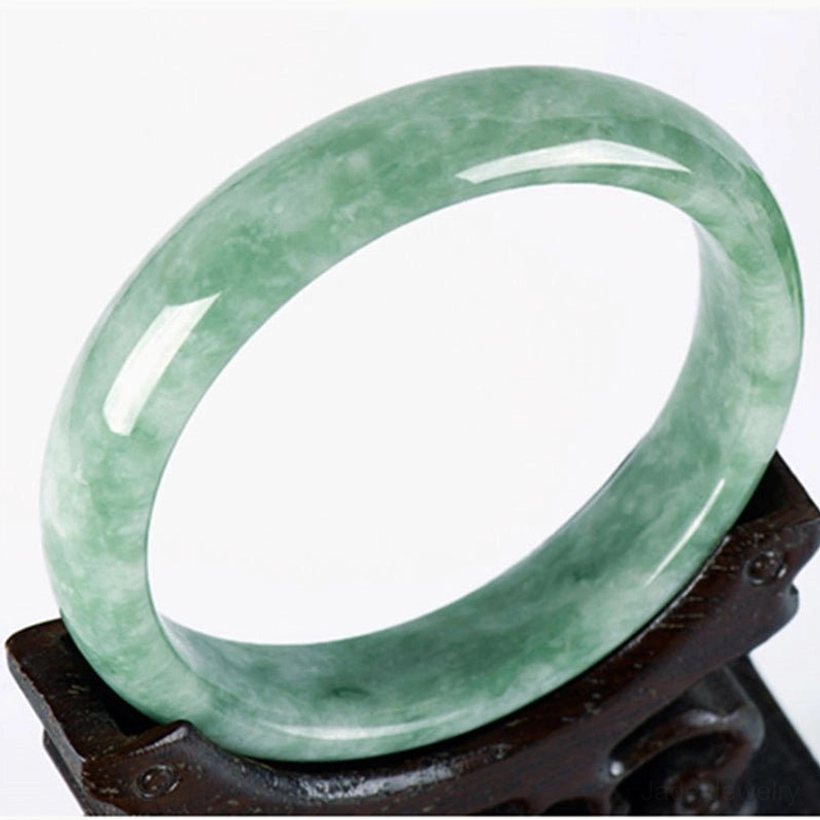 Natural jade prosperity bangle bracelet with light green mottled texture displayed on carved wooden holder, three-quarter top-down view showing authentic jade craftsmanship