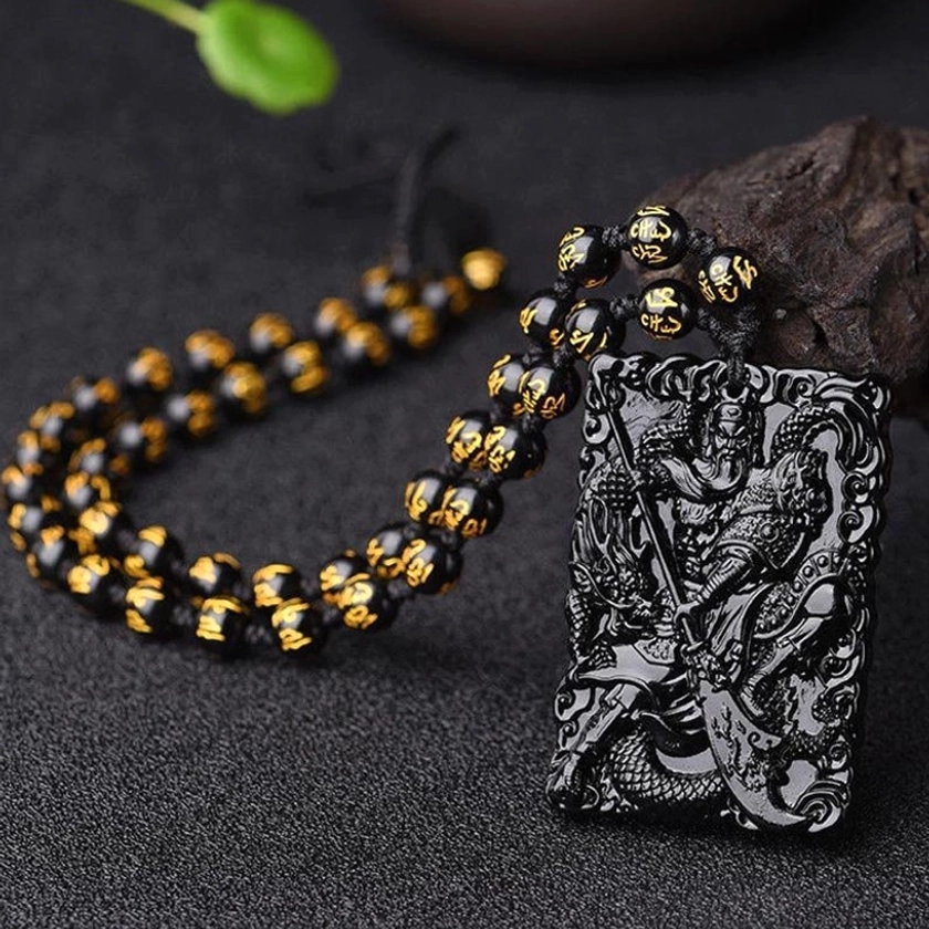 Black obsidian Lord Guan protection necklace flat lay front view with carved pendant and black-gold mantra bead chain on dark textured background
