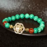 Om Mani Padme Hum jade prosperity bracelet with rotating gold charm displayed on dark ceramic plate, showcasing green jade beads, red agate accent, and crystal-adorned rose charm