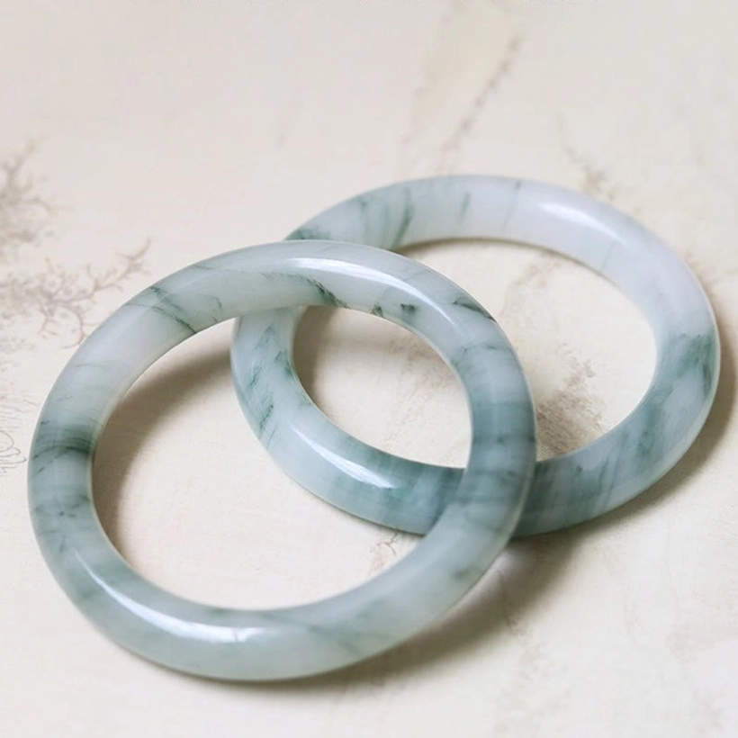 Two natural jade bangles with subtle green vein patterns displayed on textured light surface, showing polished quality and matching pair