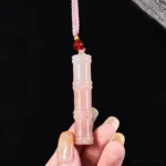Close-up hand-held view of pink quartzite jade bamboo pendant necklace, displaying carved bamboo joint details, natural stone veining, and pink braided cord with red accent bead on black background
