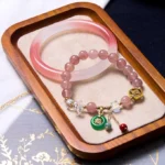 Pink jade bangle and strawberry quartz beaded bracelet set with Fu character charm, displayed on wooden tray with Chinese mat