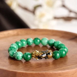 Green jade pixiu fortune bracelet with gold enamel ornament on round wooden tray, flat lay photography